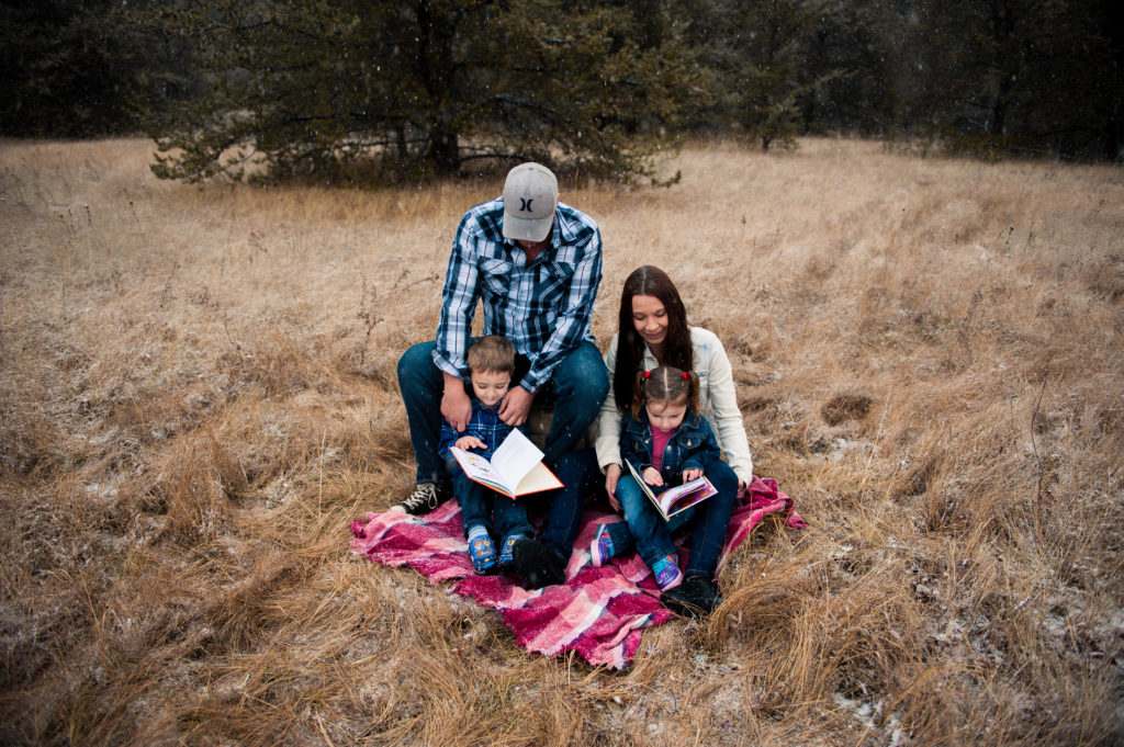 Young parents help their toddler son and daughter read Dr. Suess books in the forest, sitting on a picnic blanket.
